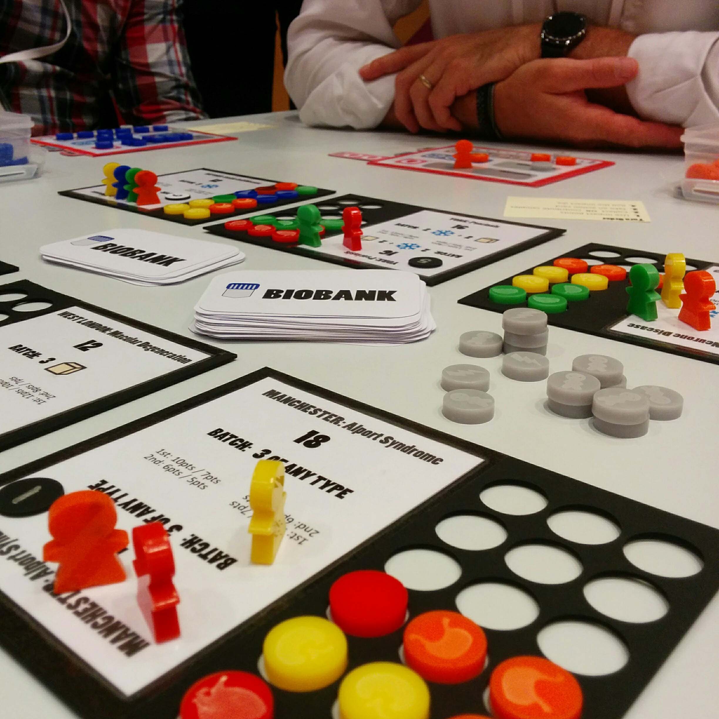 A black player board for a board game that is half filled with with red and yellow circular pieces. There are also red and yellow meeples on the board. A person's folded arms can be seen in the background.