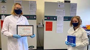 Two people stand at a distance in a lab facing the camera, both wearing face masks. One holds the Biobank of the Year Award certificate while the other holds the tropy